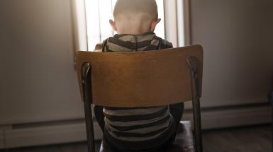 child sitting alone in a chair with back to viewer and their head is pointed downward